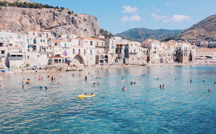 A small beach town on the side of a cliff in Sicily, Italy. People are swimming in crystal clear blue water.